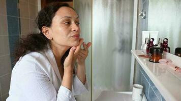 Close-up of a pregnant woman in white waffle bathrobe, applying and rubbing cleansing lotion, washing her face, standing in front of mirror in the home bathroom. Hygiene and purity concept. Skin care video