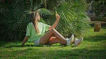 The girl takes a selfie sitting in the park on the grass in the shade video