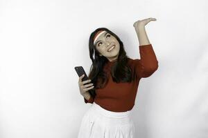 A happy Asian woman wearing headband, holding her phone, and pointing copy space on top of her, isolated by white background. Indonesia's independence day photo