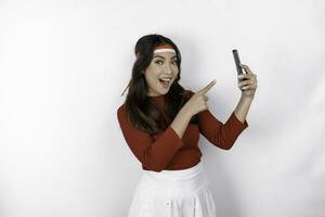 A portrait of a shocked Asian woman wearing headband, holding her phone while her mouth wide open, isolated by white background. Indonesia's independence day concept photo