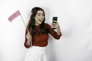 A happy Asian woman wearing headband, holding her phone and Indonesian flag, isolated by white background. Indonesia's independence day photo