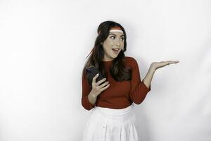 A happy Asian woman wearing headband, holding her phone, and pointing copy space beside her, isolated by white background. Indonesia's independence day photo