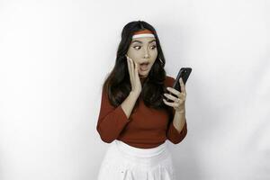 A portrait of a shocked Asian woman wearing headband, holding her phone while her mouth wide open, isolated by white background. Indonesia's independence day concept photo