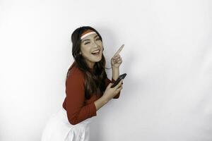 A happy Asian woman wearing headband, holding her phone, and pointing copy space beside her, isolated by white background. Indonesia's independence day photo