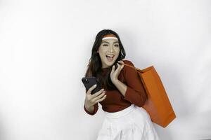 An attractive Asian woman standing excited holding an online shopping bag and her smartphone, studio shot isolated on white background. Indonesia's Independence day concept photo