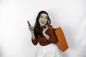 An attractive Asian woman standing excited holding an online shopping bag and her smartphone, studio shot isolated on white background. Indonesia's Independence day concept photo