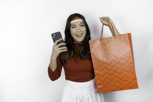 An attractive Asian woman standing excited holding an online shopping bag and her smartphone, studio shot isolated on white background. Indonesia's Independence day concept photo