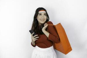 An attractive Asian woman standing excited holding an online shopping bag and her smartphone, studio shot isolated on white background. Indonesia's Independence day concept photo