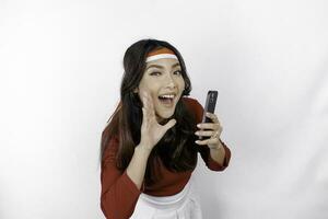 Young beautiful woman wearing a red top is holding her phone while shouting and screaming loud with a hand on her mouth. Indonesia's independence day concept. photo