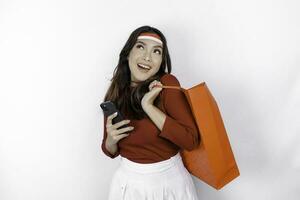 An attractive Asian woman standing excited holding an online shopping bag and her smartphone, studio shot isolated on white background. Indonesia's Independence day concept photo