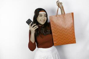 An attractive Asian woman standing excited holding an online shopping bag and her smartphone, studio shot isolated on white background. Indonesia's Independence day concept photo