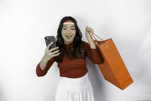 An attractive Asian woman standing excited holding an online shopping bag and her smartphone, studio shot isolated on white background. Indonesia's Independence day concept photo