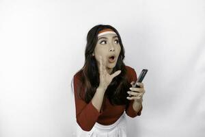 Young beautiful woman wearing a red top is holding her phone while shouting and screaming loud with a hand on her mouth. Indonesia's independence day concept. photo