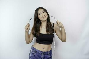 Portrait of happy young Asian woman wearing body measuring tape around waist holding spoon and fork isolated over white background photo