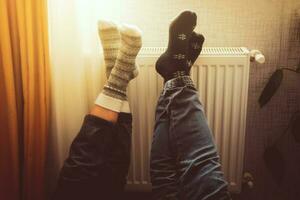 Couple hold legs up heating feet in cold home indoors on radiator in winter with cozy winter socks on. Valentines funny together warm feet by radiator by window photo