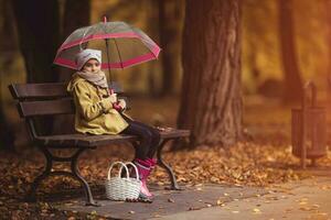 pequeño niña con un paraguas y un cesta en un parque banco foto