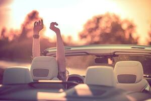 relajante en un convertible durante puesta de sol. contento joven mujer con su manos arriba. foto