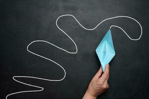 A female hand holds a paper boat against a black chalkboard background with a long path. Concept of mentorship and assistance photo