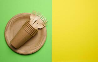 Brown paper plate, stack cups and wooden fork, disposable tableware on green background, top view. Copy space photo