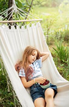 A little girl rests in a hammock and eats cherries in the summer. photo
