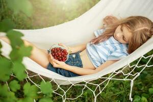 A little girl rests in a hammock and eats cherries in the summer. photo