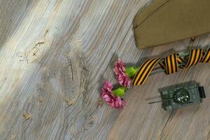 Two pink carnations, Saint George ribbon, tank, and military cap on a wooden surface. photo