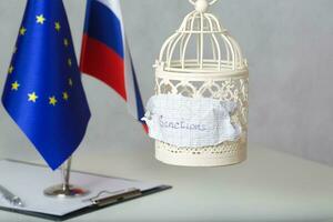 The small cage and the word SANCTIONS attached to it. Flags of EU and Russia in the background photo