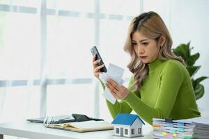 Young Asian woman sitting and calculating expenses and mortgage with calculator and house on table Worried about payments and non-repayable loans, business, and property. photo