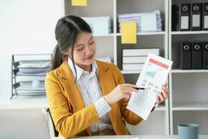 Asian businesswoman worker or manager in a yellow suit in a good mood using a laptop and calculator to calculate company income and expenses balance inside the office. photo