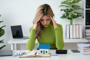 oung Asian woman sitting and calculating expenses and mortgage with calculator and house on table Worried about payments and non-repayable loans, business, and property photo