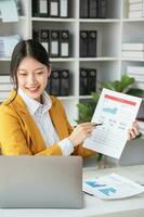 asiático mujer de negocios trabajador o gerente en un amarillo traje en un bueno estado animico utilizando un ordenador portátil y calculadora a calcular empresa ingresos y gastos equilibrar dentro el oficina. foto