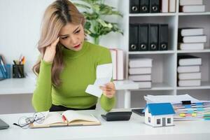 oung Asian woman sitting and calculating expenses and mortgage with calculator and house on table Worried about payments and non-repayable loans, business, and property photo