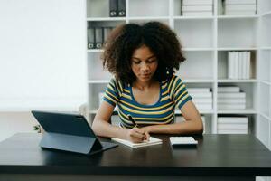 Beautiful African woman using laptop and tablet while sitting at her working place. Concentrated at work in home office photo