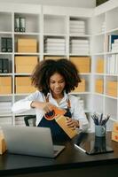 puesta en marcha pequeño negocio pequeño, empresario propietario africano mujer utilizando teléfono inteligente o tableta tomando recibir y comprobación en línea compra compras orden a preparando paquete producto caja. foto