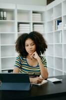 hermosa africano mujer utilizando ordenador portátil y tableta mientras sentado a su trabajando lugar. concentrado a trabajo en hogar oficina foto
