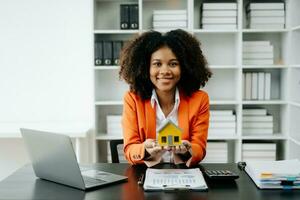 Young real estate agent worker working with laptop and tablet at table in office and small house beside it. photo