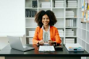 Young real estate agent worker working with laptop and tablet at table in office and small house beside it. photo