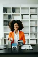 Young real estate agent worker working with laptop and tablet at table in office and small house beside it. photo