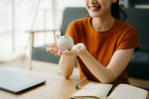 asiático De las mujeres mano ahorro un moneda dentro cerdito banco con de madera a calcular y tableta en el mesa para negocio, finanzas, ahorro dinero y propiedad inversión concepto. foto