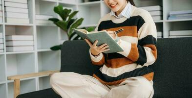 Young Asian woman listening music from headphones and writing note for her work idea in diary book.She in living room on the sofa at home photo