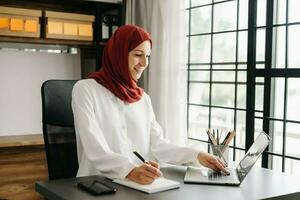 joven Arábica hembra empresario vistiendo un hijab trabajando en línea con un ordenador portátil a oficina foto