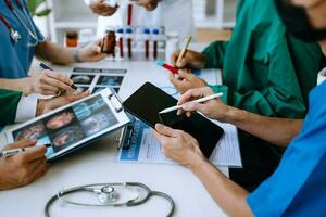 Medical team having a meeting with doctors in white lab coats and surgical scrubs seated at a table discussing a patients working online using computers photo