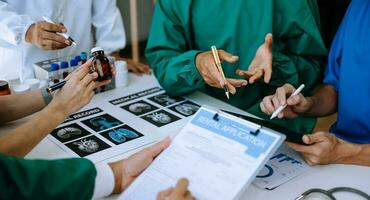 Medical team having a meeting with doctors in white lab coats and surgical scrubs seated at a table discussing a patients working online using computers photo