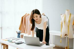 Asian young woman on desk in office of fashion designer and holds tablet, laptop and smartphone on white table photo