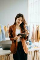 Moda diseñador mujer hablando inteligente teléfono y utilizando ordenador portátil con digital tableta computadora en moderno estudio el ropa colgando en el bastidores Mañana ligero foto