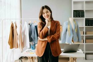 asiático Sastre mujer trabajando en ropa en sastrería taller. hermosa joven hembra Moda diseñador sonrisa y después éxito en moderno estudio foto