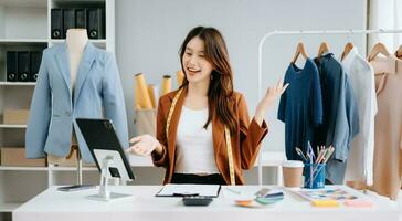 Moda diseñador mujer hablando inteligente teléfono y utilizando ordenador portátil con digital tableta computadora en moderno estudio el ropa colgando en el bastidores Mañana ligero foto