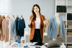 Fashion designer woman talking smart phone and using laptop with digital tablet computer in modern studio the clothes hanging on the racks morning light photo