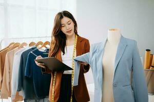 Moda diseñador mujer hablando inteligente teléfono y utilizando ordenador portátil con digital tableta computadora en moderno estudio el ropa colgando en el bastidores Mañana ligero foto