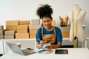 puesta en marcha pequeño negocio pequeño, empresario propietario africano mujer utilizando teléfono inteligente o tableta tomando recibir y comprobación en línea compra compras orden a preparando paquete producto caja. en oficina foto